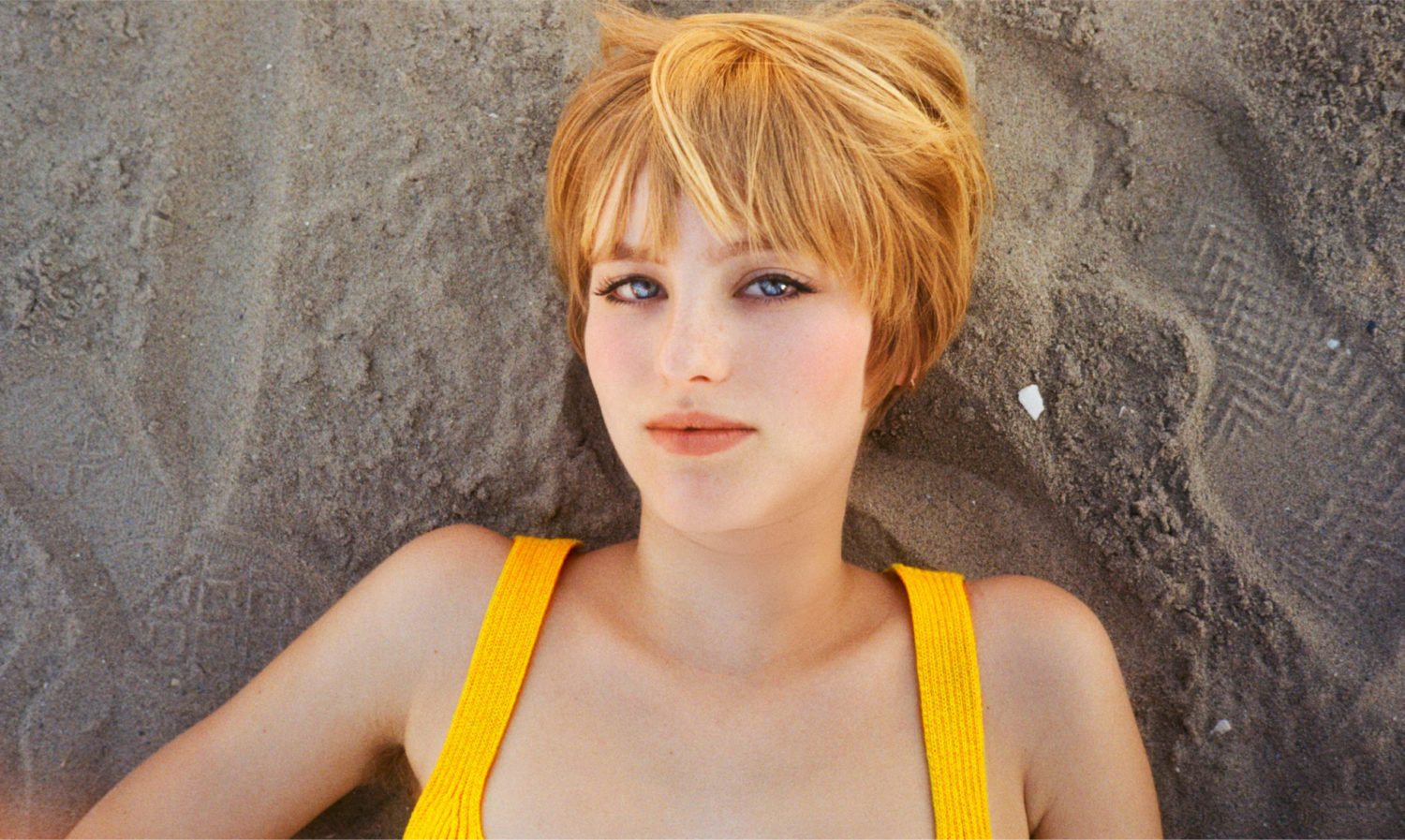 A woman with a yellow tank top and blue eyes is lying on the sand